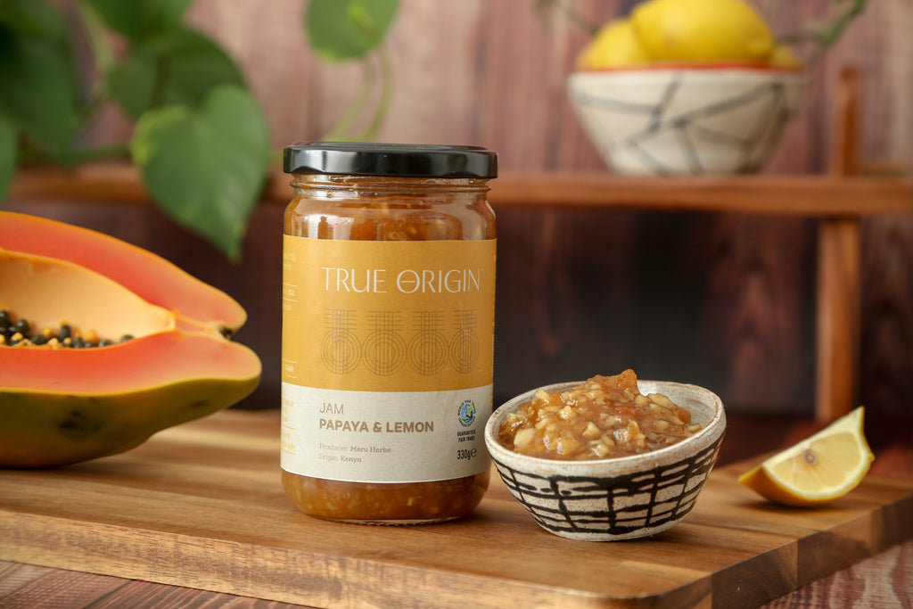 A glass jar of Papaya & Lemon Jam with a label that reads 'TRUE ORIGIN' and 'PAPAYA & LEMON JAM'. The jar is sealed with a black lid.  It is sitting on a wooden chopping board next to a bowl filled with papaya and lemon jam and a cut open fresh papaya and a bowl of lemons in the background.