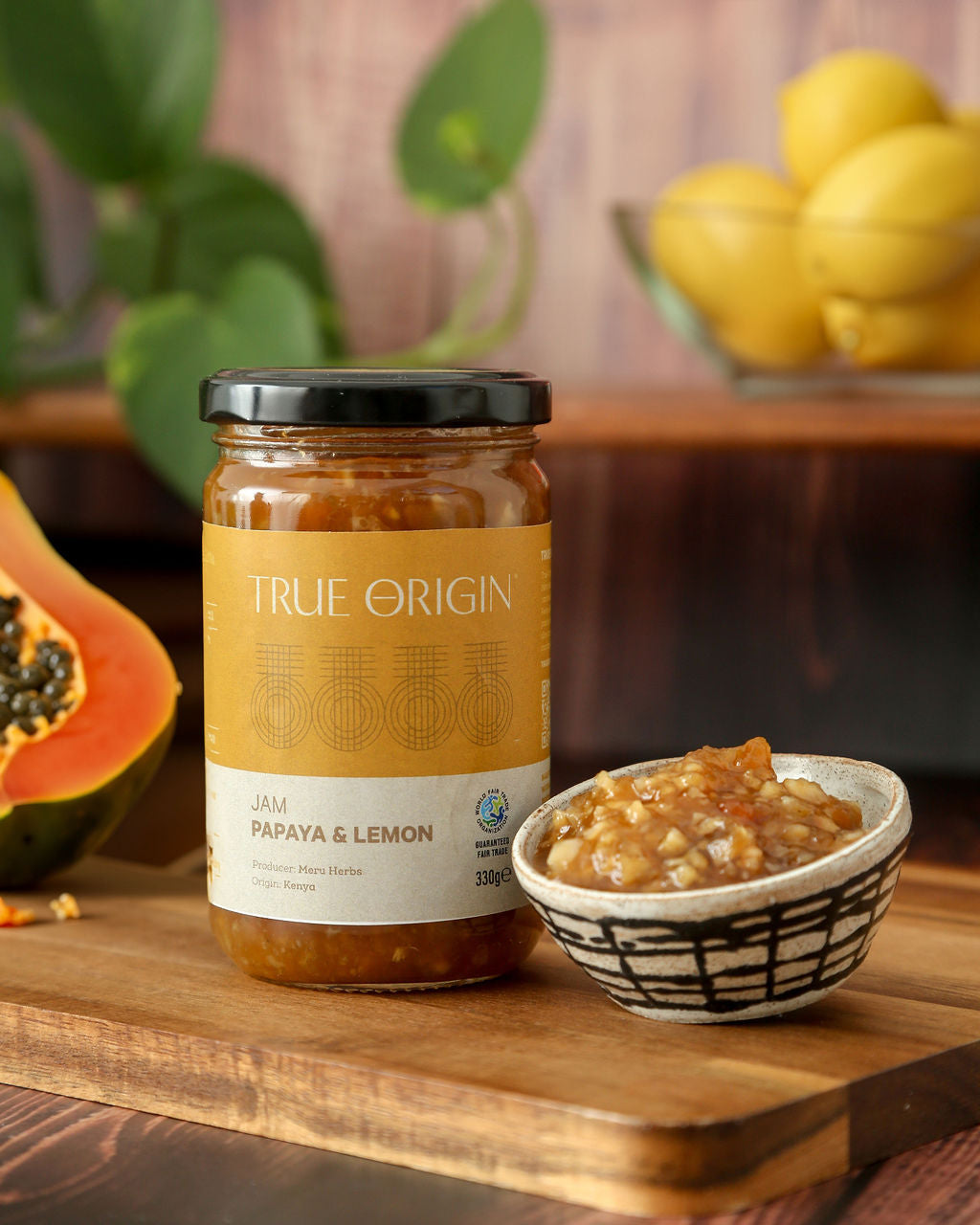 A jar of True Origin papaya and lemon jam on a wooden chopping board with an open papaya and a bowl with papaya jam in it.  There is a bowl of lemons in a bowl in the background.