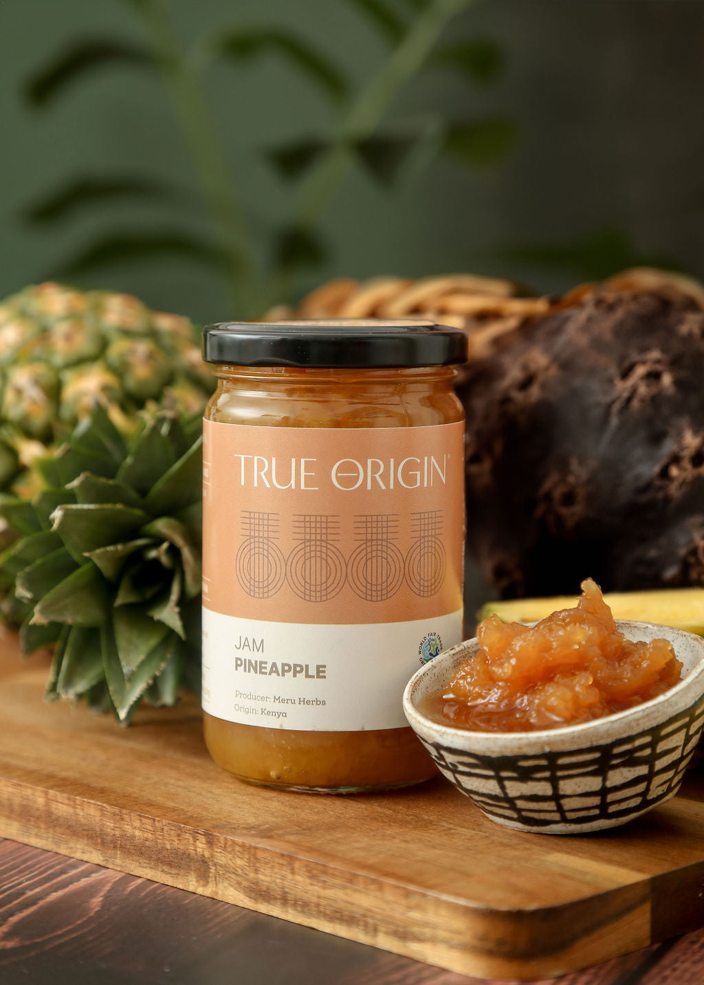 A jar of True Origin Pineapple Jam with an orange and white label next to a fresh pineapple and a bowl of pineapple jam.