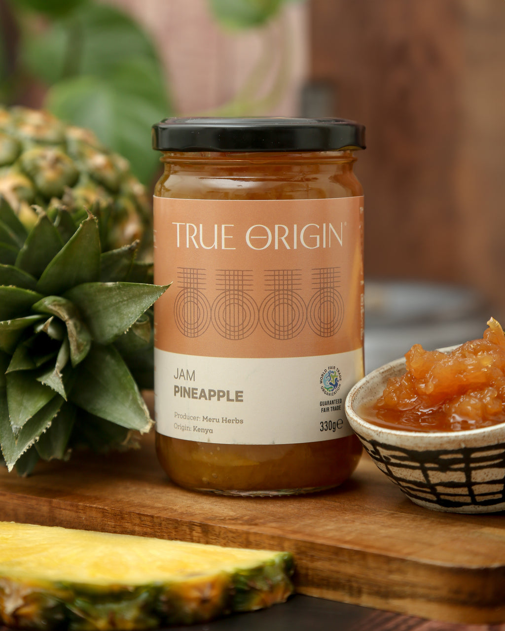 A glass jar of Pineapple Jam with the label 'TRUE ORIGIN' and 'Pineapple' written on it, indicating the flavour of the jam.  It is sitting next to a bowl filled with the jam and a fresh pineapple.