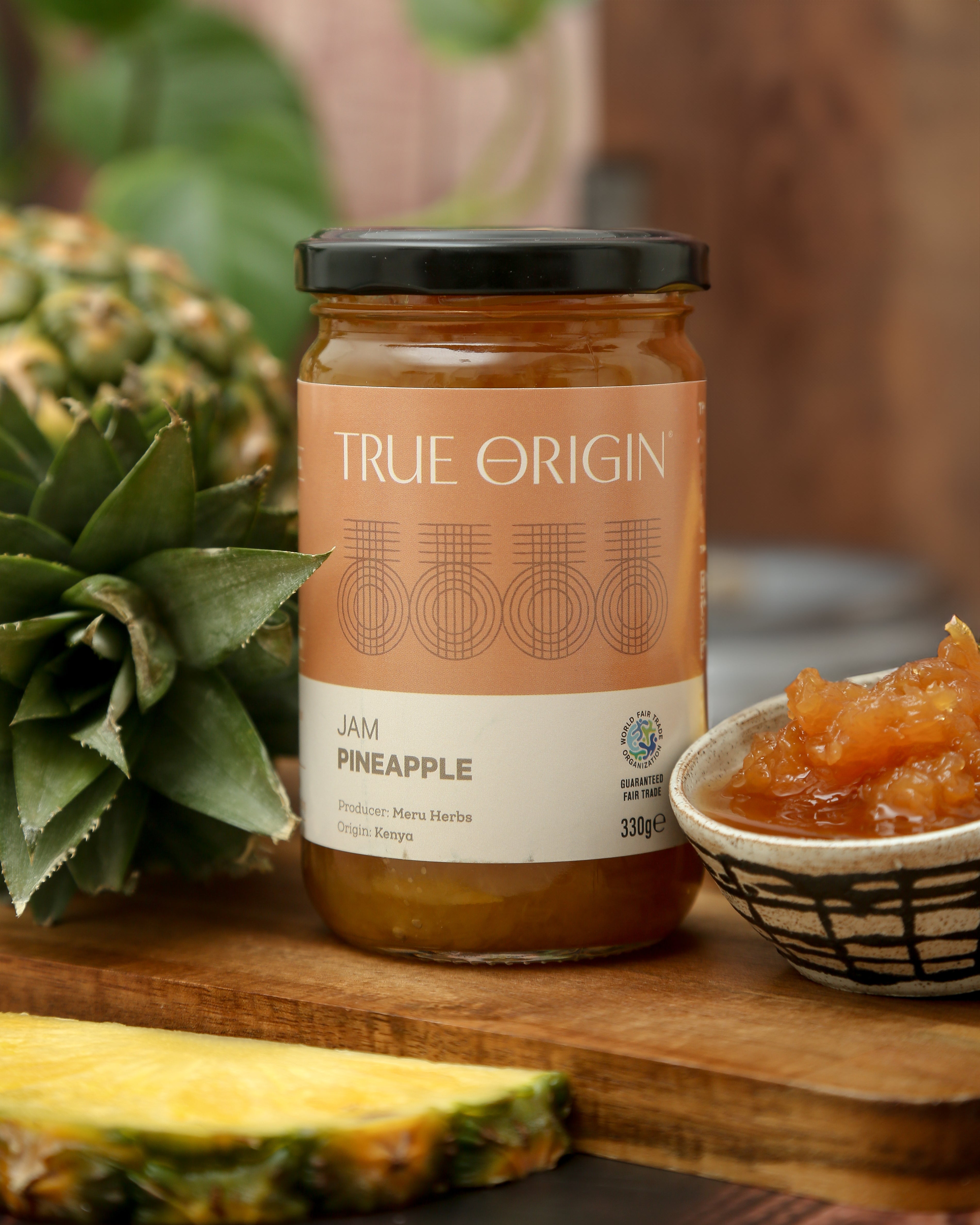 A glass jar of Pineapple Jam with the label 'TRUE ORIGIN' and 'Pineapple' written on it, indicating the flavour of the jam.  It is sitting next to a bowl filled with the jam and a fresh pineapple.