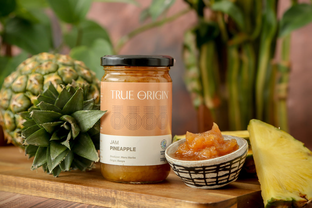 A glass jar of Pineapple Jam with the label 'TRUE ORIGIN' and 'Pineapple' written on it, indicating the flavour of the jam.  It is sitting next to a bowl filled with the jam and a fresh pineapple.