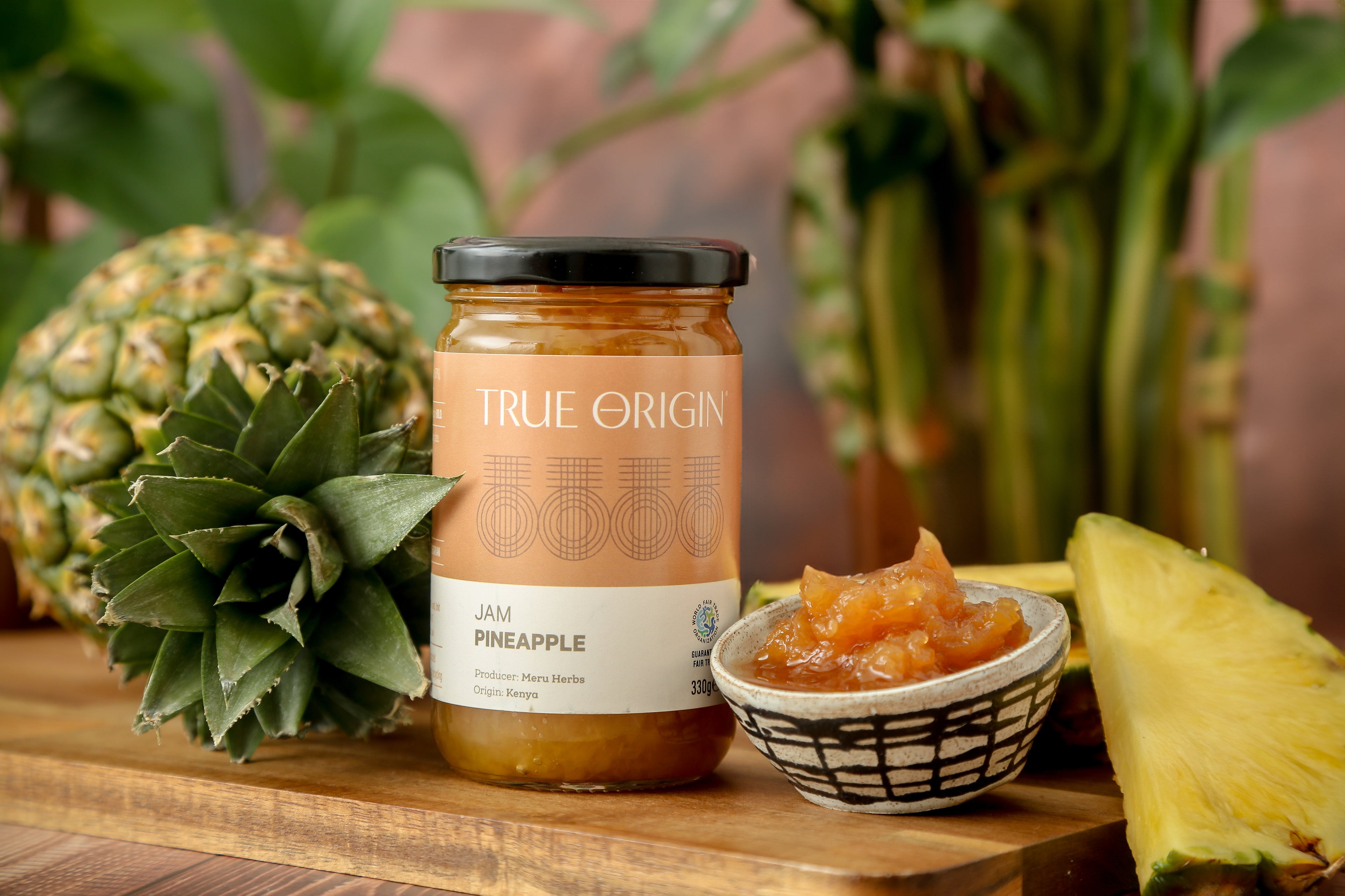 A glass jar of Pineapple Jam with the label 'TRUE ORIGIN' and 'Pineapple' written on it, indicating the flavour of the jam.  It is sitting next to a bowl filled with the jam and a fresh pineapple.