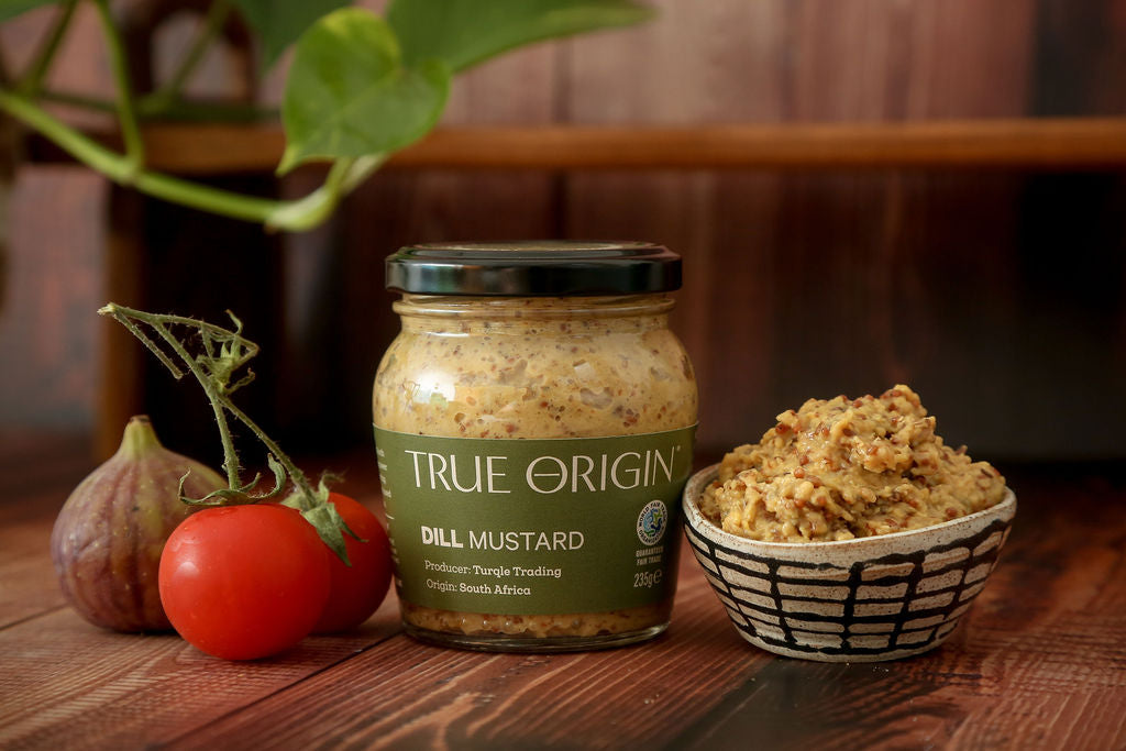 Jar of True Origin dill mustard with a small bowl of mustard and vegetables on a wooden surface.