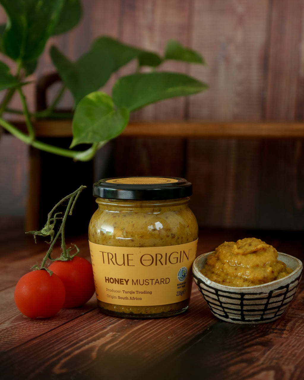 A glass jar of Honey Mustard branded by True Origin, with a label indicating the product is produced by Turqle Trading, origin South Africa, and the weight of 235g.  There is a bowl filled with Honey mustard and some fresh tomatoes
