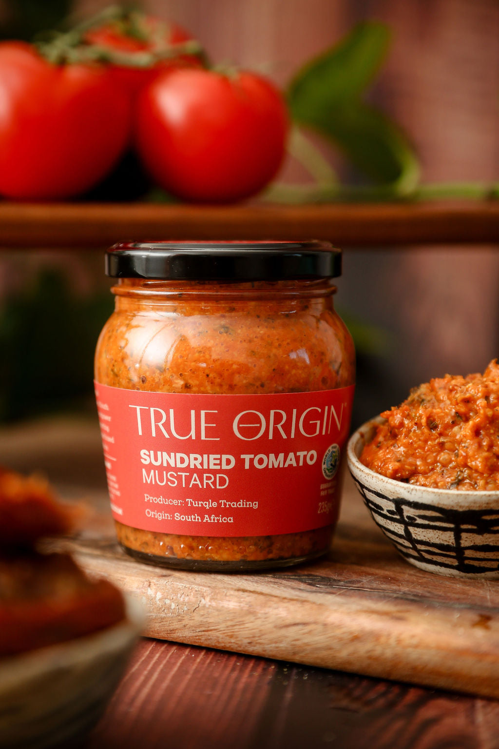 A red jar of True Origin Sundried Tomato Mustard beside the mustard in a bowl and some tomatoes in the background.