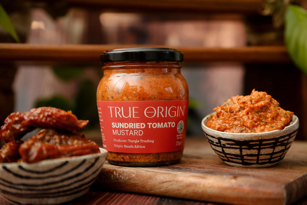 An unopened red jar of True Origin Sundried Tomato Mustard on a wooden chopping board beside a bowl of sundried tomatoes and a bowl of sundried tomato mustard.