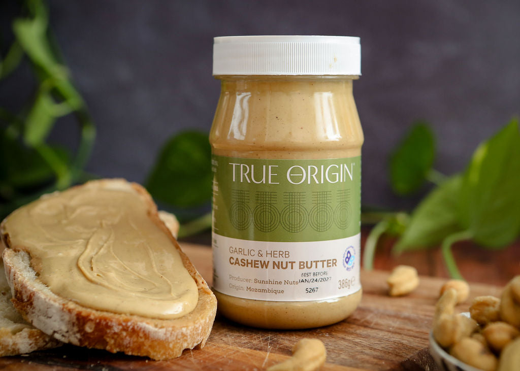 Jar of True Origin cashew nut butter on a wooden board with bread and nuts.
