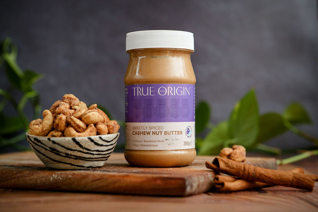 Jar of True Origin cashew nut butter with cashews and cinnamon sticks on a wooden surface.