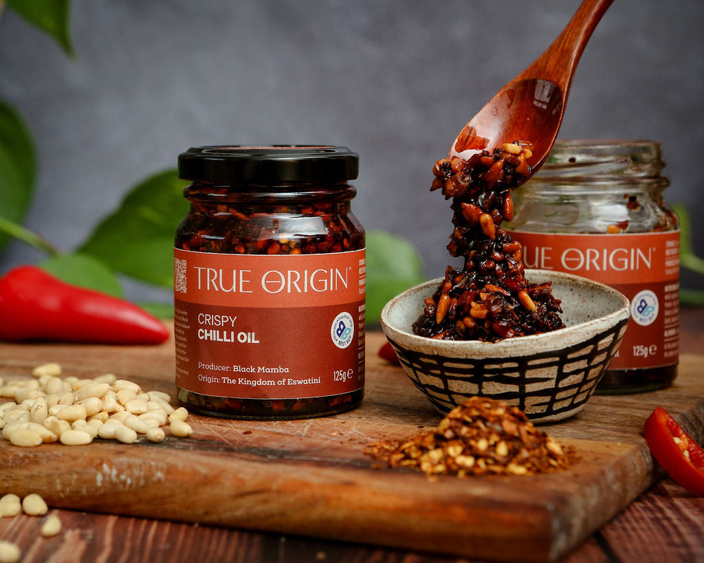 Jar of True Origin Crispy Chilli Oil with a bowl of contents coming from a spoon, sitting on a wooden chopping board.
