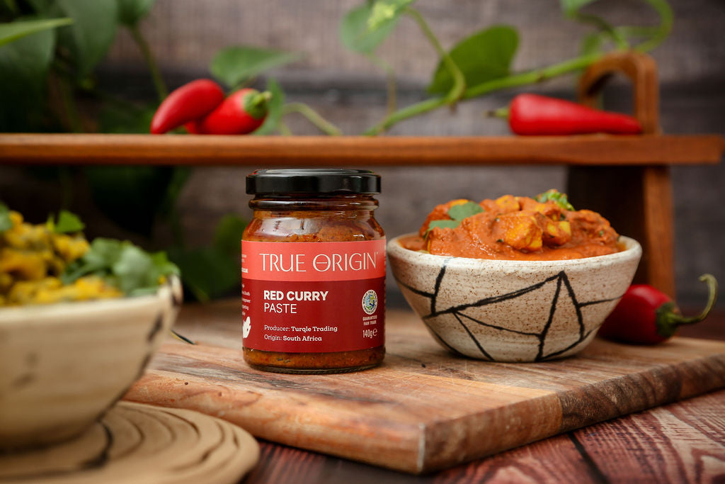 An unopened red jar of True Origin Red Curry Paste on a wooden chopping board next to a bowl of red curry and some chillies on a shelf in the background.