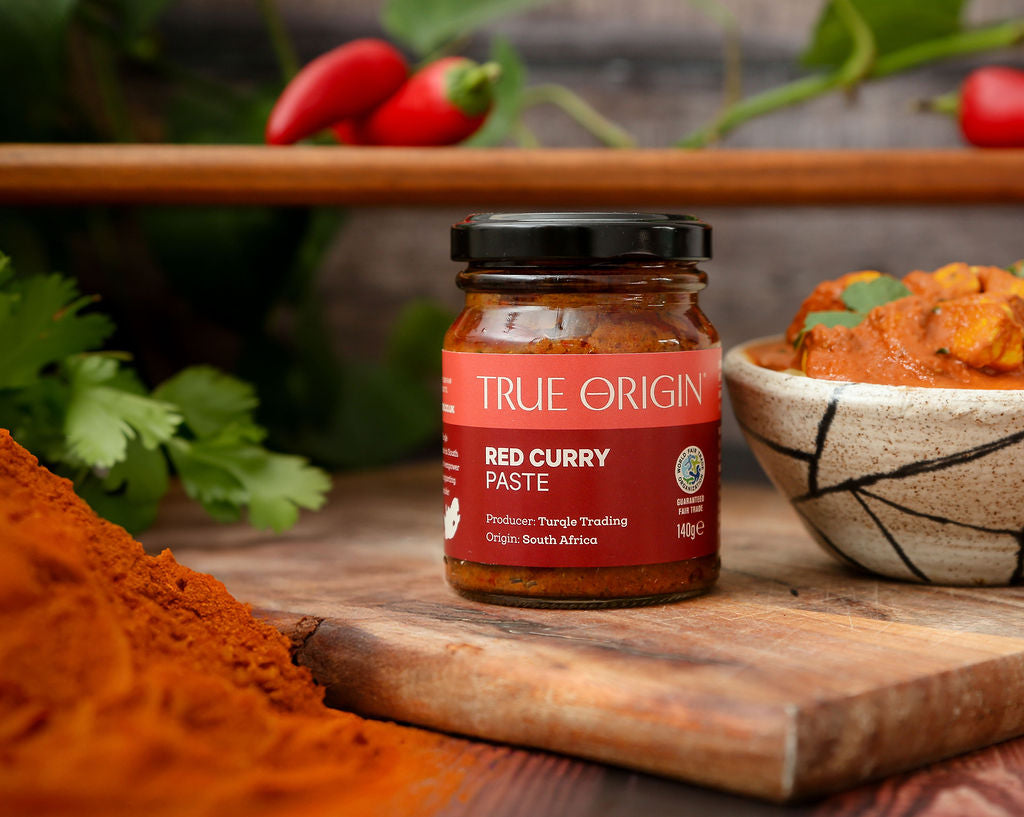 An unopened red jar of True Origin Red Curry Paste on a wooden chopping board beside a bowl of red curry and a pile of curry powder.