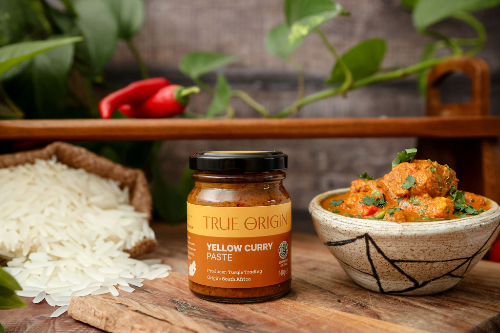 An unopened yellow jar of True Origin Yellow Curry Paste on a wooden surface next to a bowl of yellow curry and some white rice with some chillies in the background.