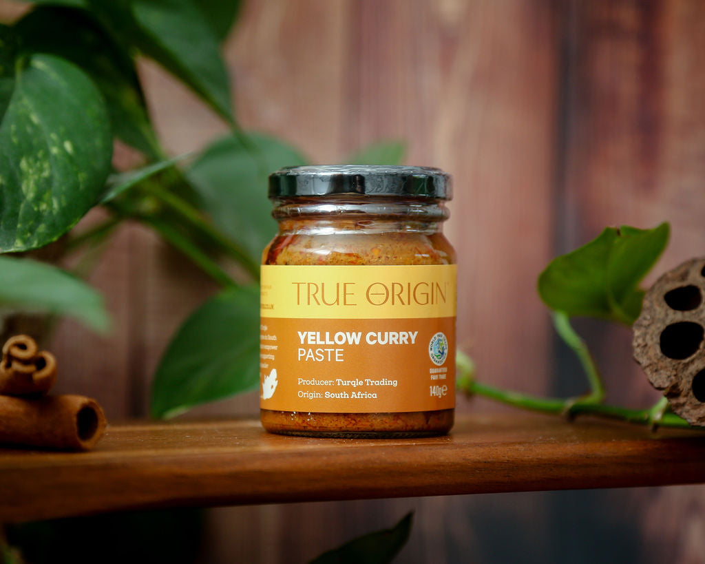 An unopened yellow jar of True Origin Yellow Curry Paste on a wooden shelf with some greenery in the background.