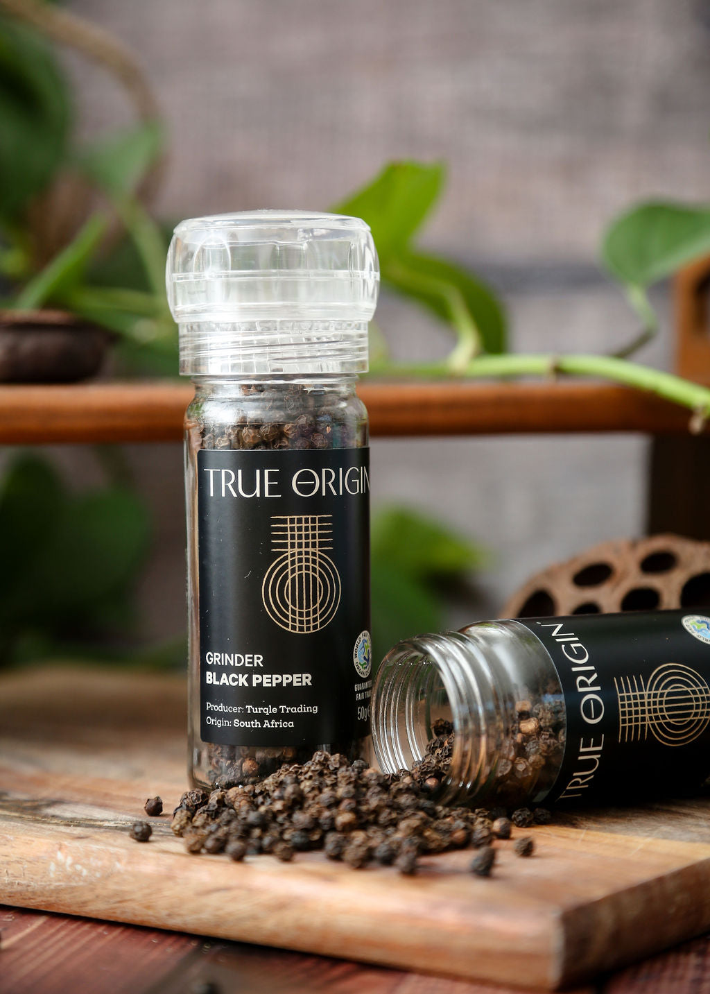 A jar of true origin black pepper with a built in grinder next to an open jar lying on its side with the black peppercorns coming out onto  a wooden chopping board.