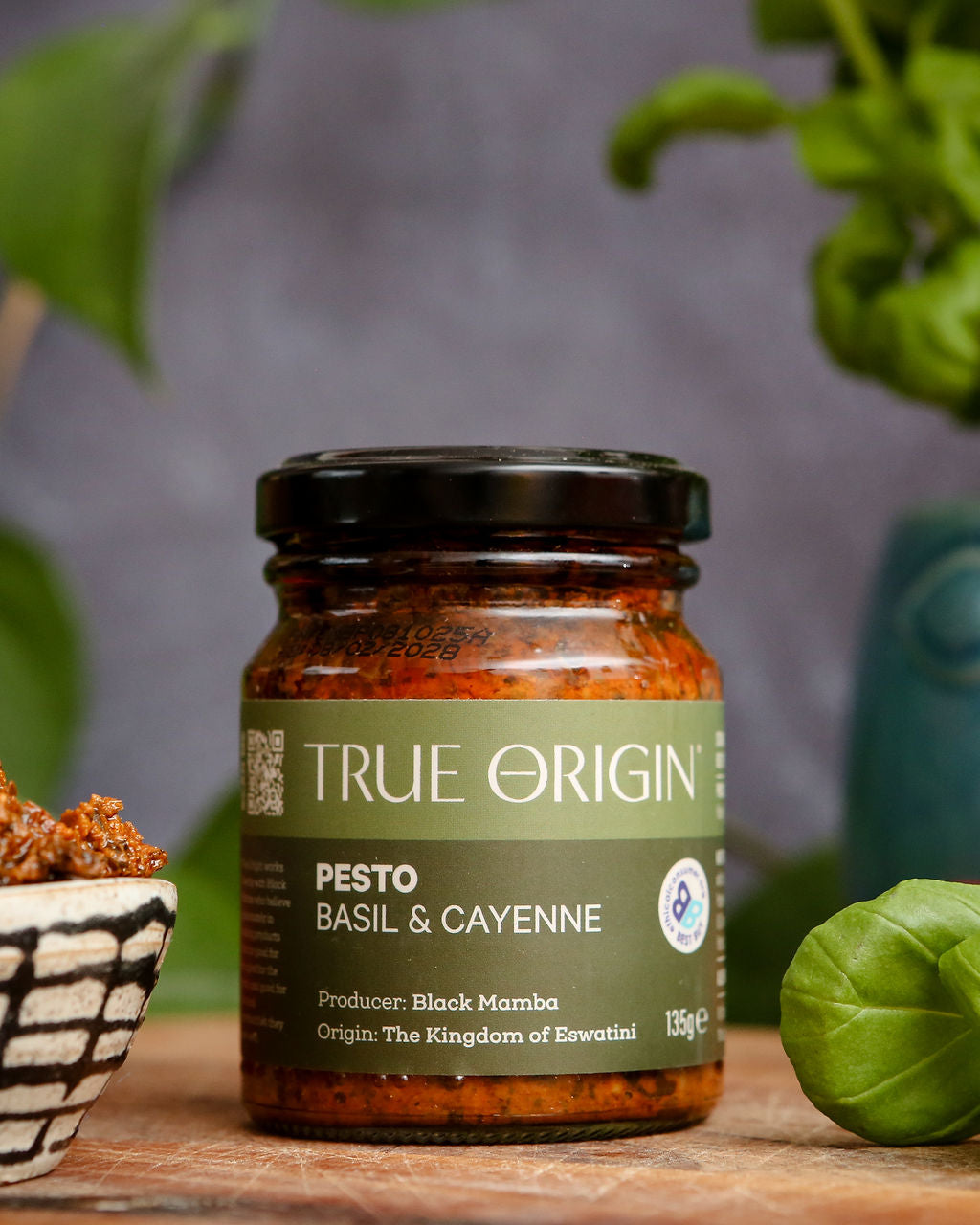 Jar of True Origin basil & cayenne pesto on a wooden surface with greenery in the background