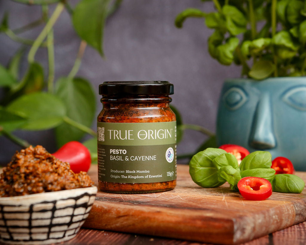 Jar of True Origin basil and cayenne pesto on a wooden cutting board with fresh basil and chillies and a bowl of contents.