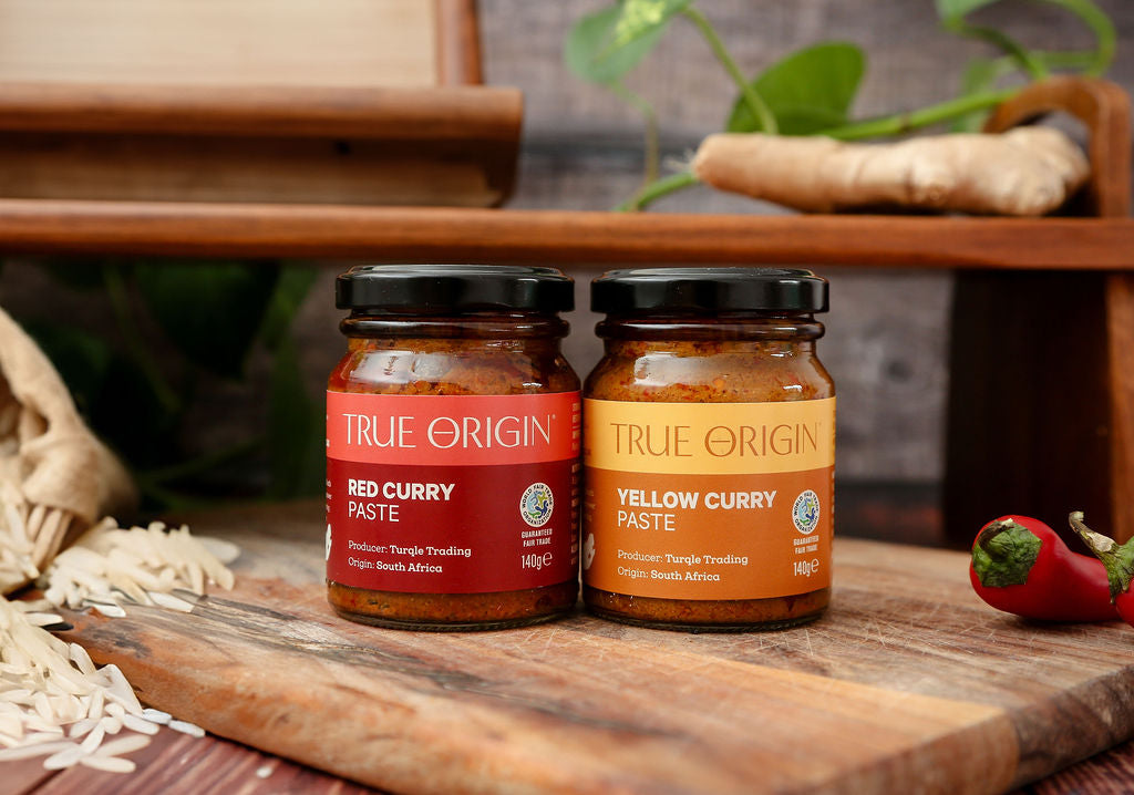 Two jars of True Origin curry paste on a wooden surface with ingredients in the background.