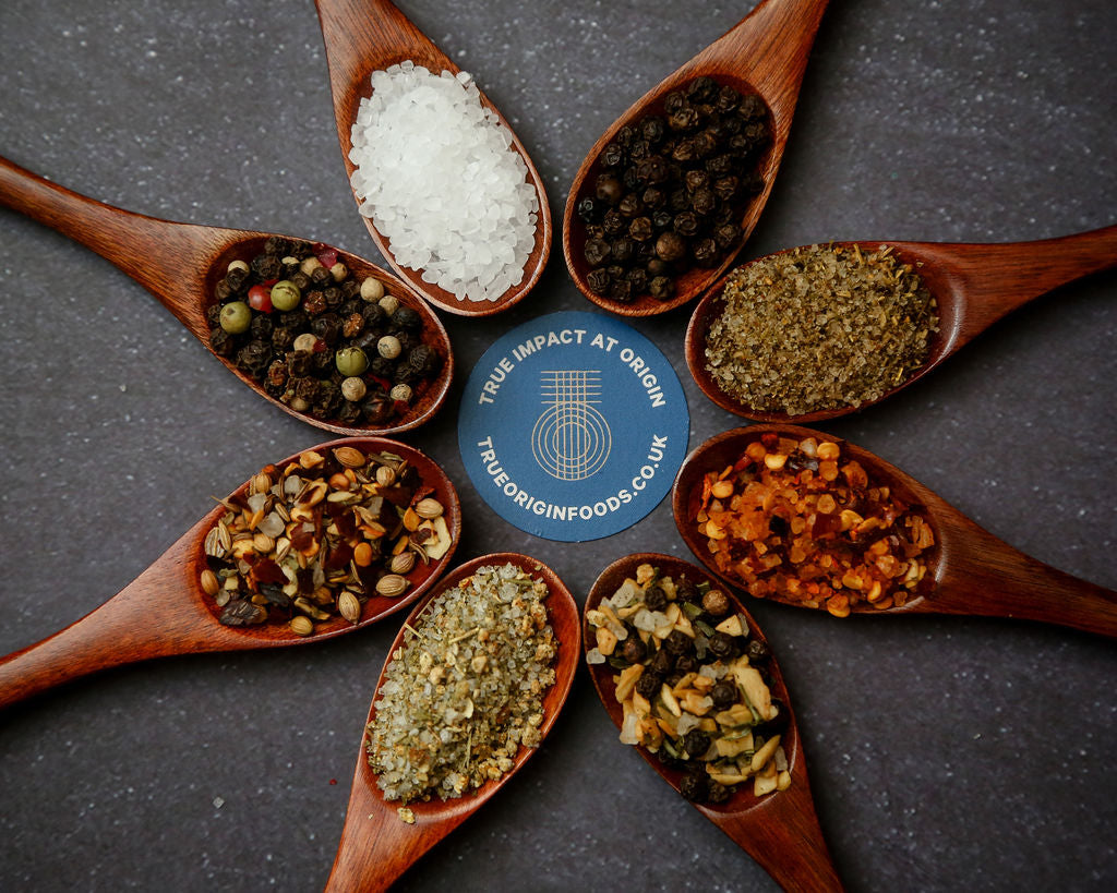 Eight wooden spoons in a circle each containing a different True Origin seasoning:  Black Pepper, Tomato Salt, Chilli Salt, Lemon Pepper, Lemon Salt, Harissa Spice, Rainbow Pepper, Khoisan Natural Salt.  In the middle is a blue circle with the True Origin Logo and the words True Impact at Origin.