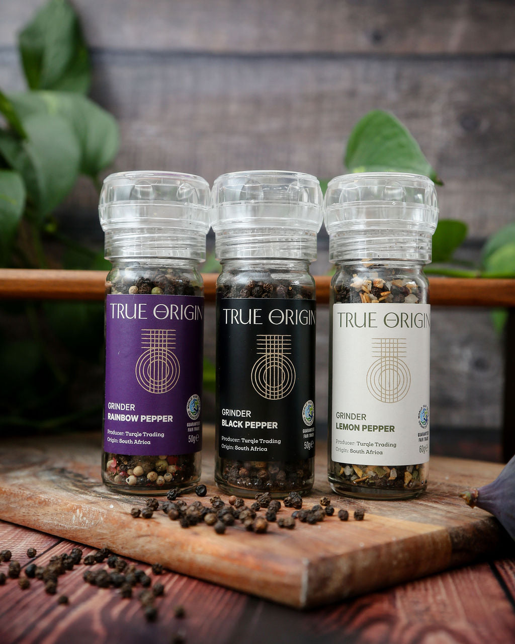 Three glass bottles with build in grinders Rainbow Pepper, Black Pepper and Lemon Pepper are in a row on a wooden chopping board with some loose peppercorns.