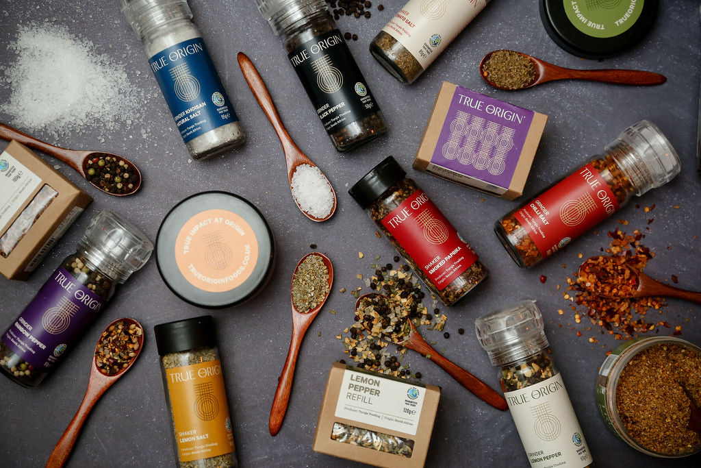 Several True Origin seasonings, grinders and shakers lying at angles on a worktop with wooden spoons containing the salts and peppercorns.