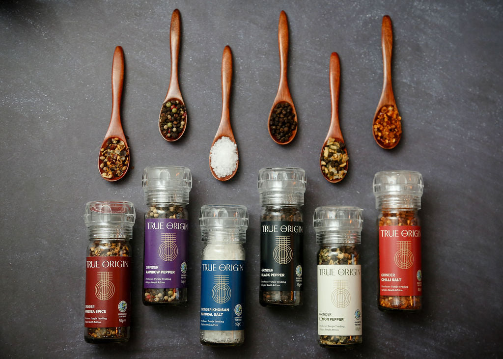 Five True Origin grinders staggered in a row below wooden spoons containing the matching ingredients. L-R there is Harissa Spice, Rainbow Pepper, Khoisan Natural Salt, Black Pepper, Lemon Pepper, Chilli Salt.