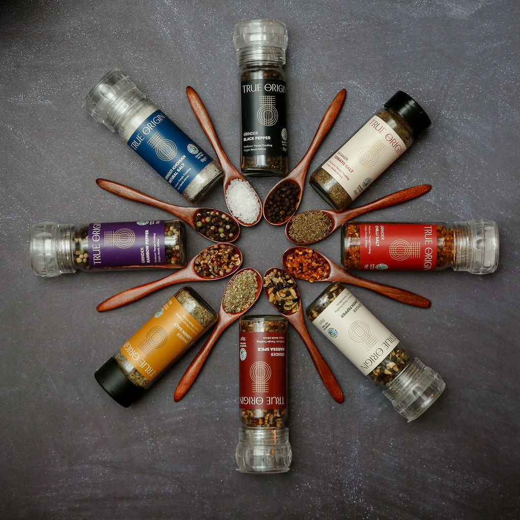 A range of eight grinders and shakers in a circle with spoons in the middle containing the seasonings.  Features True Origin Black Pepper, Tomato Salt, Chilli Salt, Lemon Pepper, Harissa Spice, Lemon Salt, Rainbow Pepper, Khoisan Natural Salt.