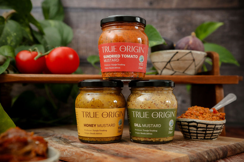 A beautiful image of three True Origin mustards with some ingredients in the background on a wooden surface and some greenery.