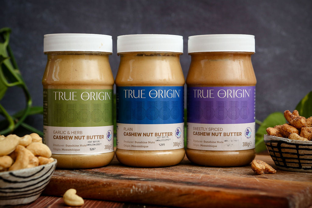 Three jars of True Origin cashew nut butter on a wooden surface with a dark background.