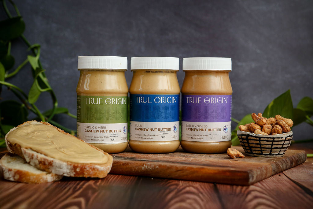 Three jars of True Origin cashew nut butter on a wooden board with bread and nuts.