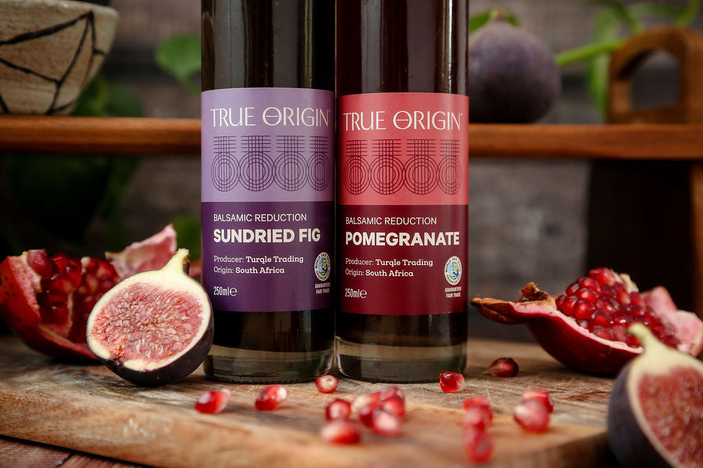 Close up image of the labels on the True Origin Sundried Fig and Balsamic Reduction bottles on a wooden chopping board with cut figs and pomegranates.