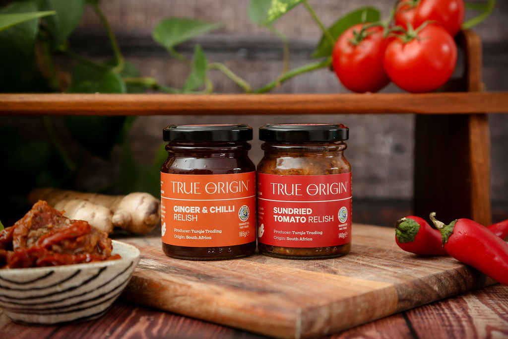 Two jars of True Origin relish on a wooden cutting board with fresh ingredients.