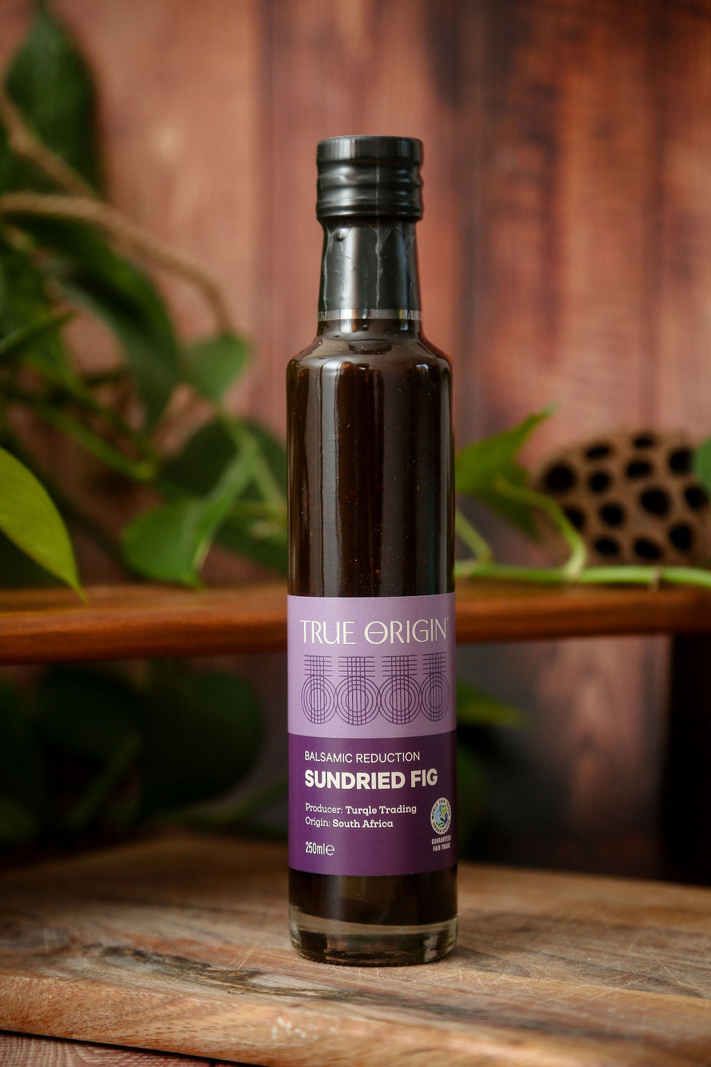 Purple bottle of Sundried Fig Balsamic Reduction on a wooden chopping board with a plant and a shelf in the background.
