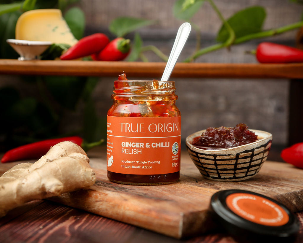 An orange jar of True Origin Ginger and Chilli relish next to some relish in a bowl and some ginger with some chillies in the background.