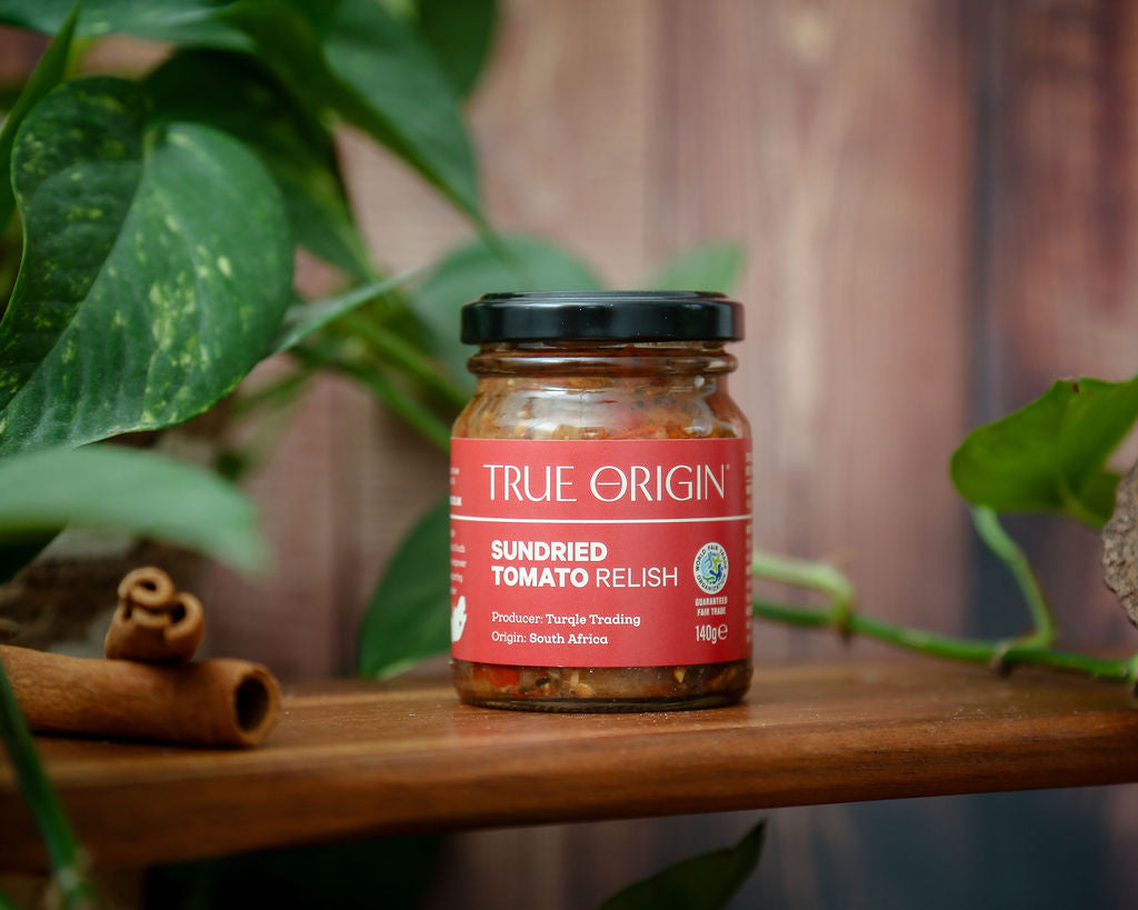 A red jar of True Origin Sundried Tomato relish with some green leaves in the background.
