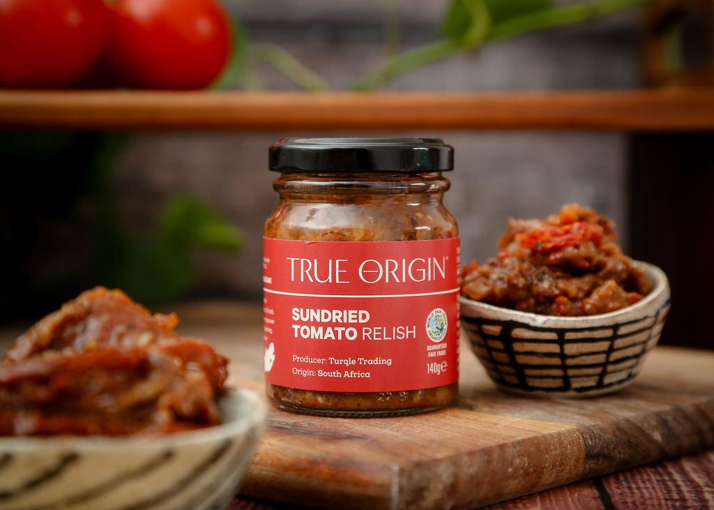 A red jar of True Origin Sundried Tomato relish next to some relish in a bowl and some tomatoes in the background.