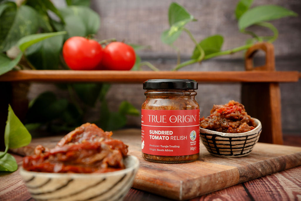 A red jar of True Origin Sundried Tomato relish next to some relish in a bowl and some tomatoes in the background.