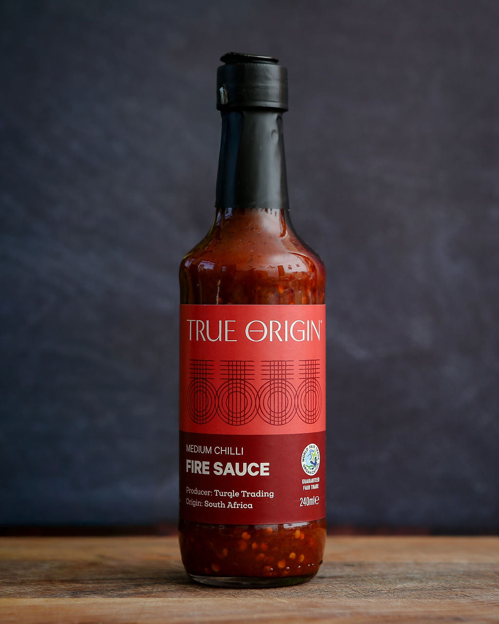 Bottle of True Origin fire sauce on a wooden surface with a dark background