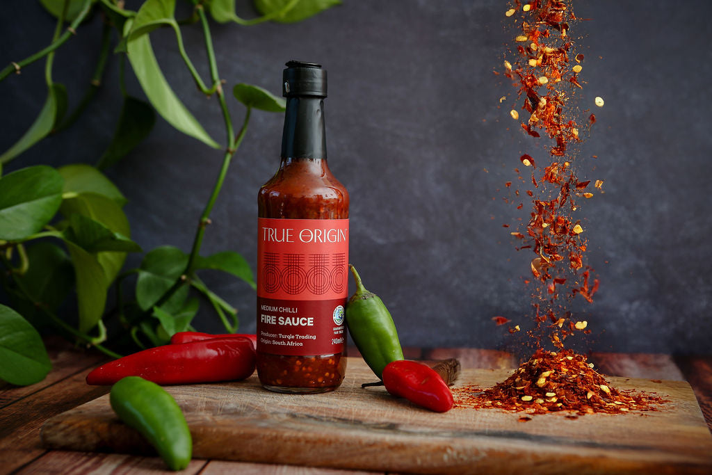 Bottle of True Origin hot sauce with chili flakes being sprinkled on a wooden board with peppers.