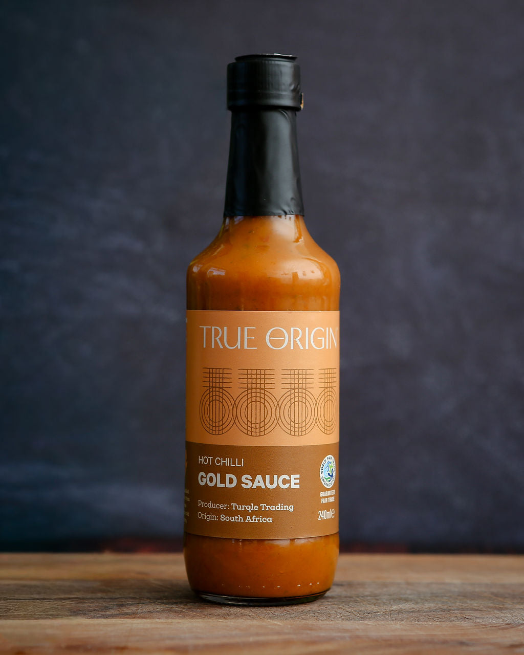 Bottle of True Origin Gold Hot Chilli Sauce on a wooden surface with a dark background