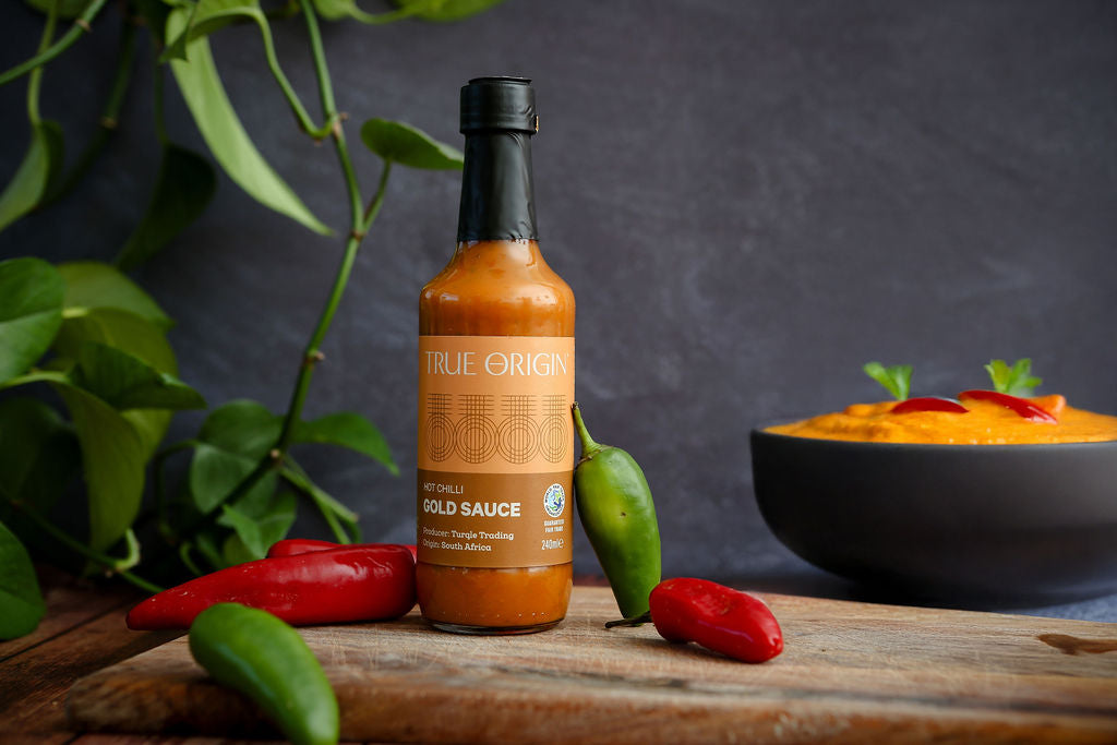 Bottle of True Origin hot sauce on a wooden surface with peppers and a bowl of food in the background.