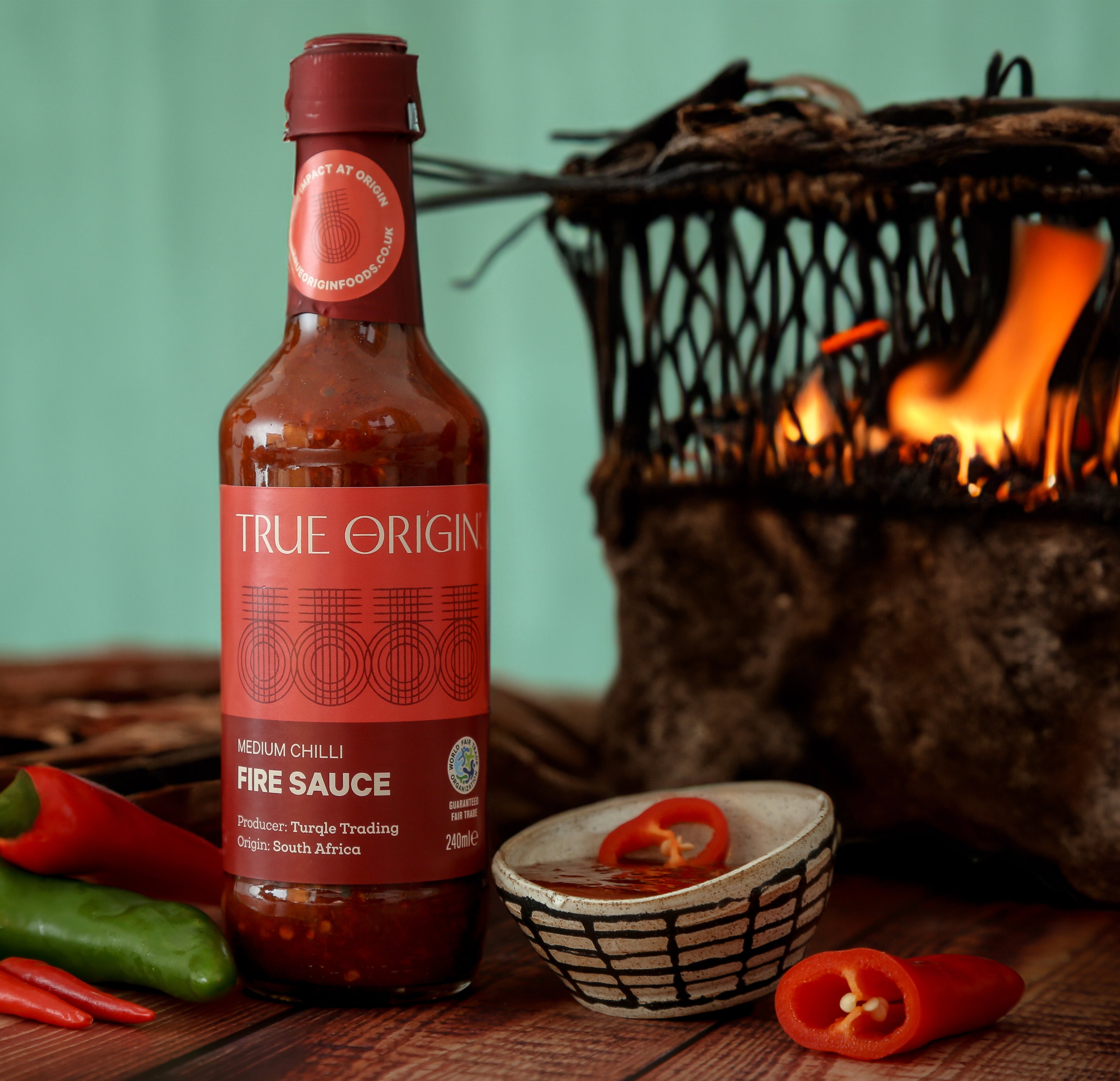 A bottle of True Origin Medium Chilli Fire Sauce with medium chilli, indicating medium spiciness level.  It is next to some fresh chillies and a fire in the background.