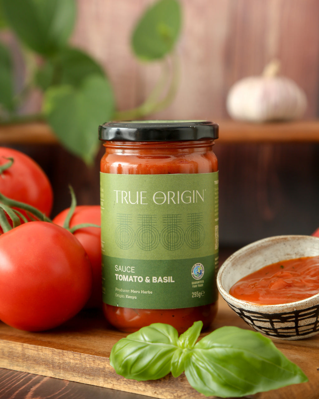 A green jar of fairtrade Tomato and Basil sauce in True Origin branding sitting on a wooden chopping board next to some fresh tomatoes and basil and a bowl containing the sauce.