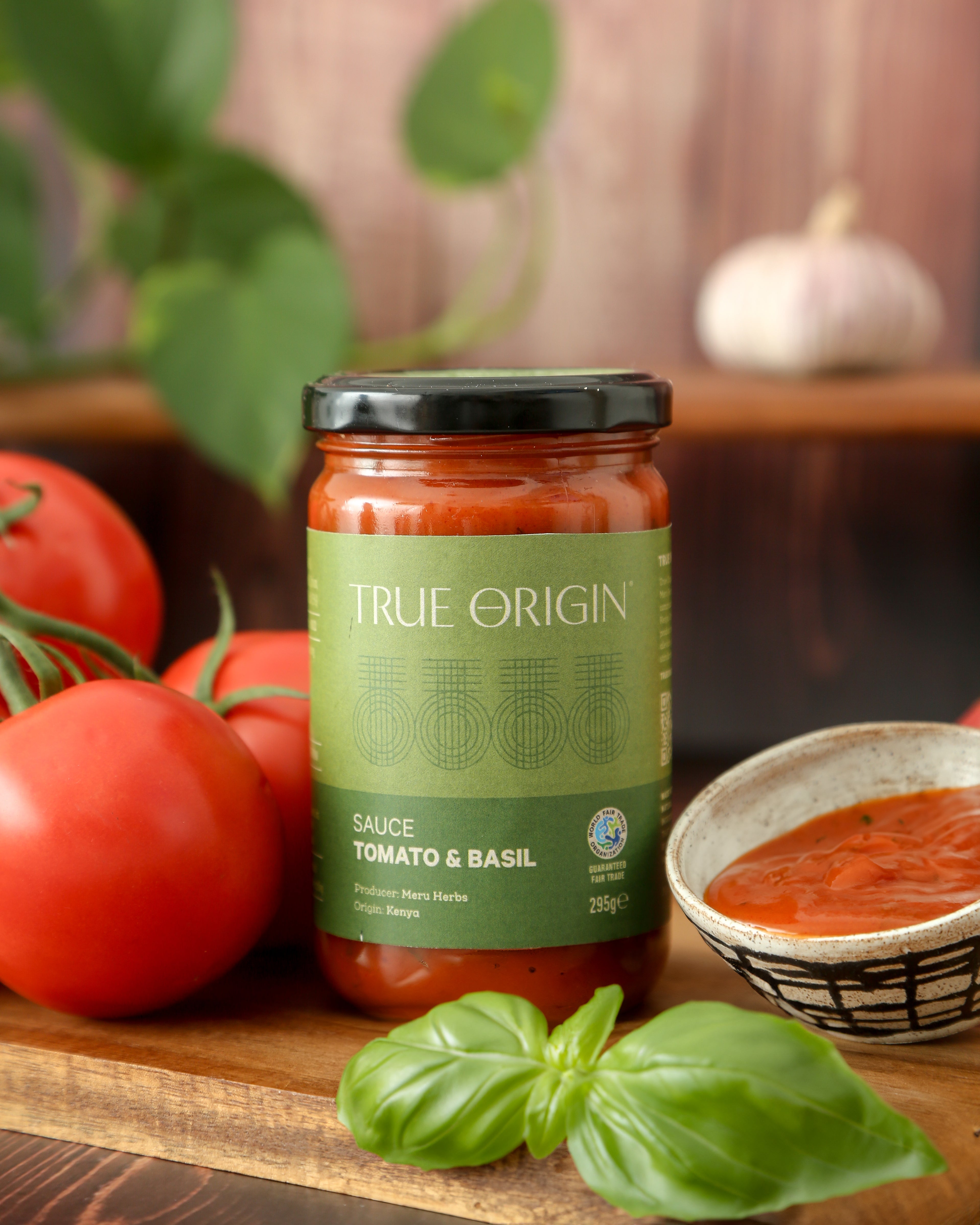 A green jar of fairtrade Tomato and Basil sauce in True Origin branding sitting on a wooden chopping board next to some fresh tomatoes and basil and a bowl containing the sauce.