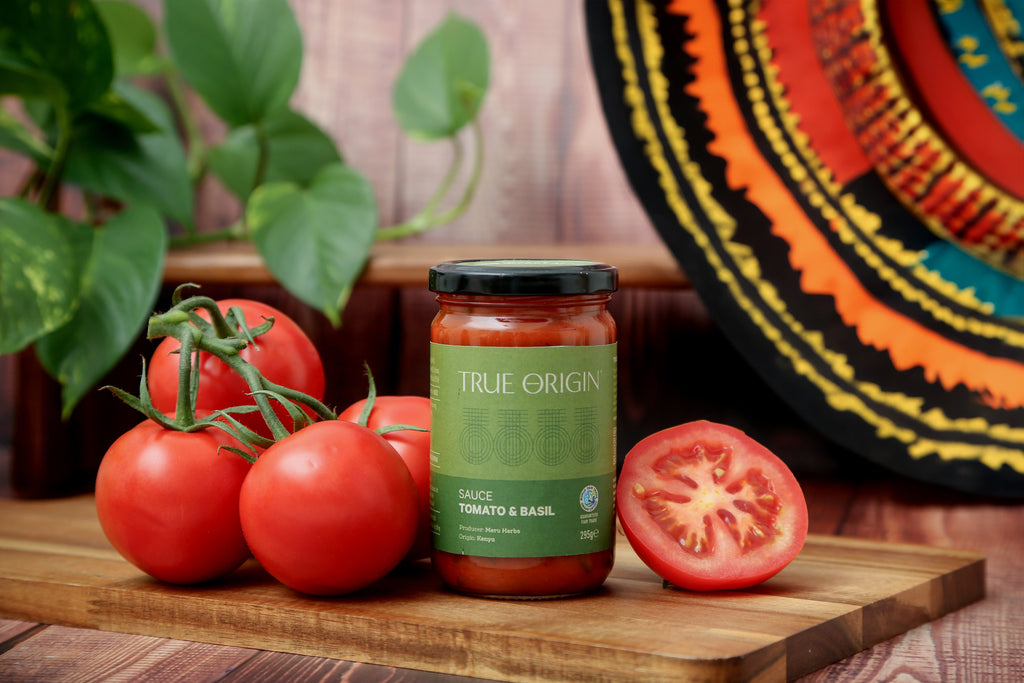 A green jar of fairtrade Tomato and Basil sauce in True Origin branding sitting on a wooden chopping board next to some fresh tomatoes.