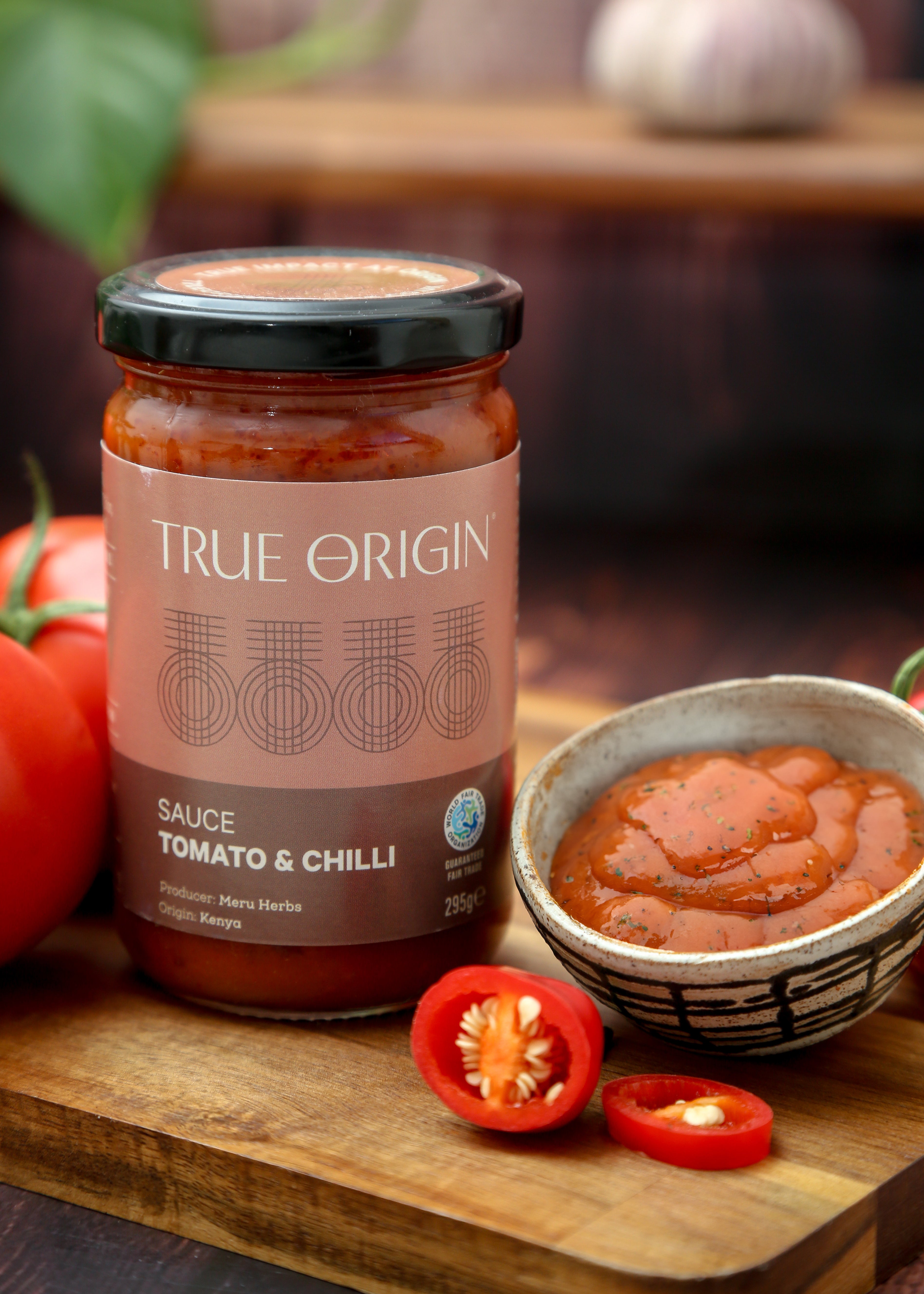 A jar of True Origin brand Tomato & Chilli sauce produced by Meru Herbs in Kenya.  The jar is sitting on a wooden chopping board next to some fresh tomatoes and a bowl containing that tomato and chilli sauce.