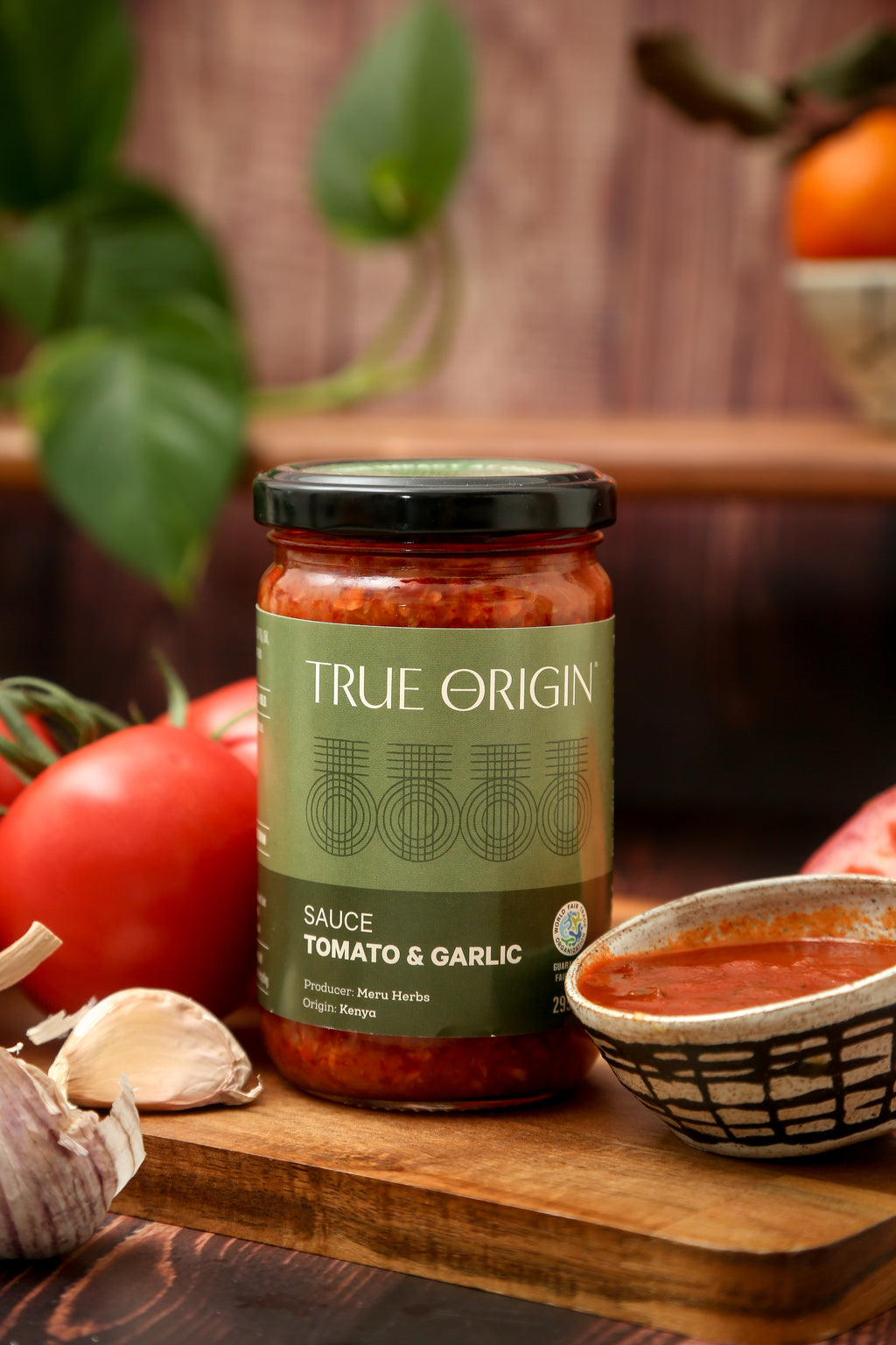 A jar of True Origin Tomato and Garlic sauce sitting on a wooden surface with a bowl of tomato and garlic sauce and some fresh tomatoes and garlic.