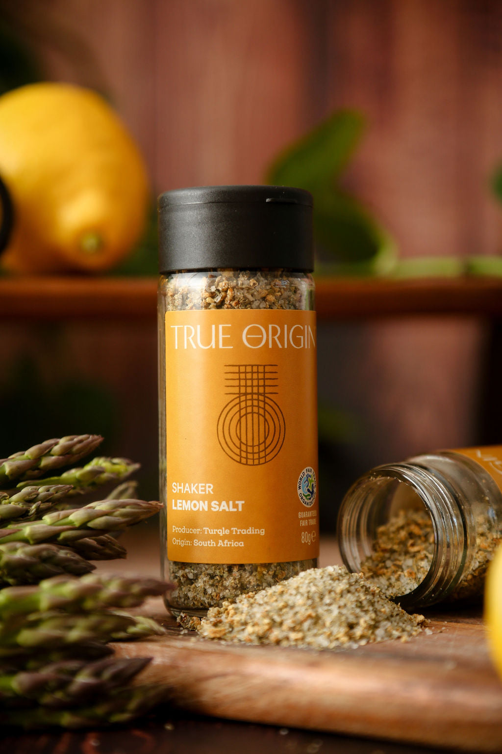 A yellow glass jar of lemon salt with a black lid and an open jar on its side with lemon salt pouring out onto a chopping board next to some asparagus and a lemon in the background.