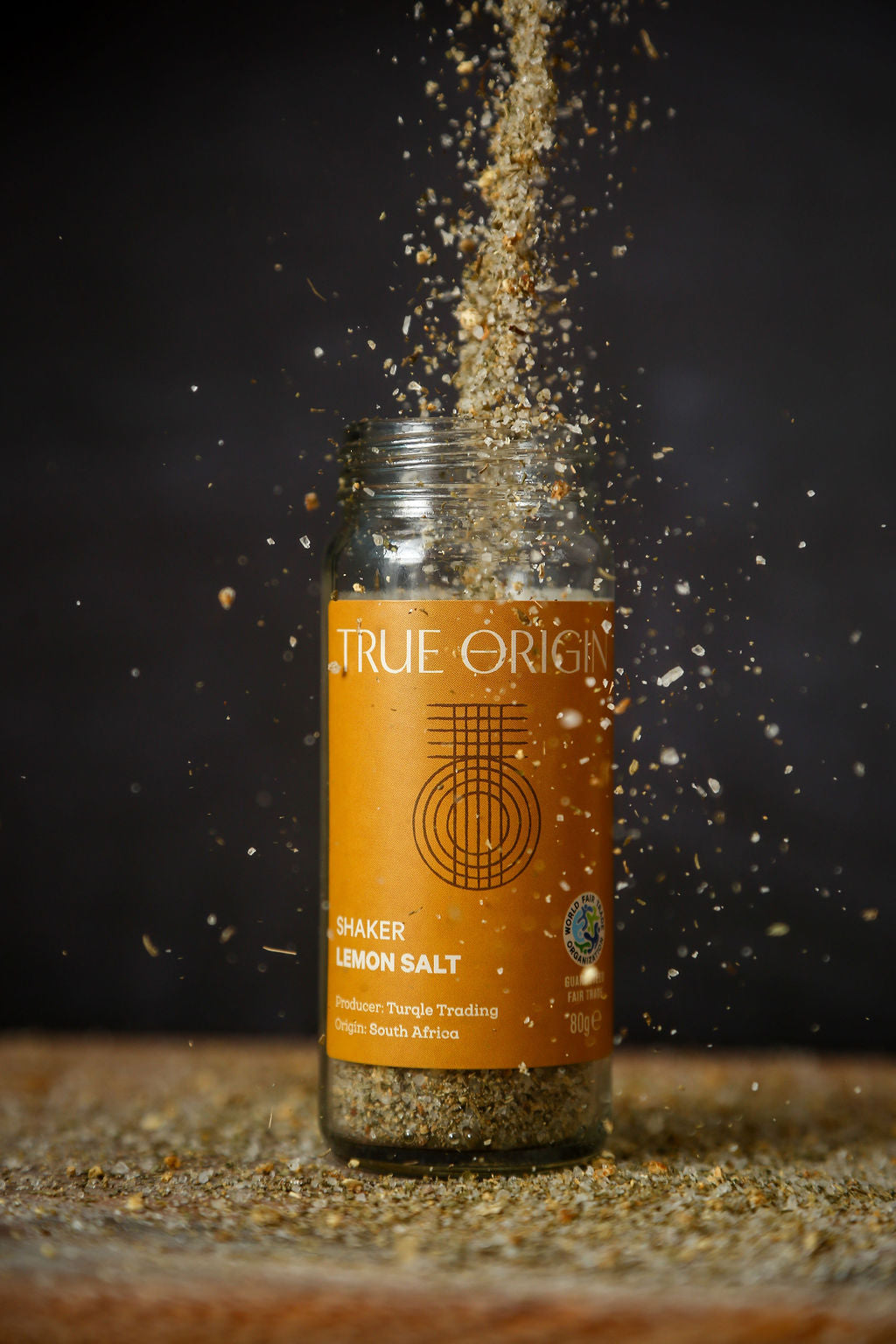 An open yellow glass jar of lemon salt with lemon salt being poured into the jar from above and sprinkling onto a chopping board.