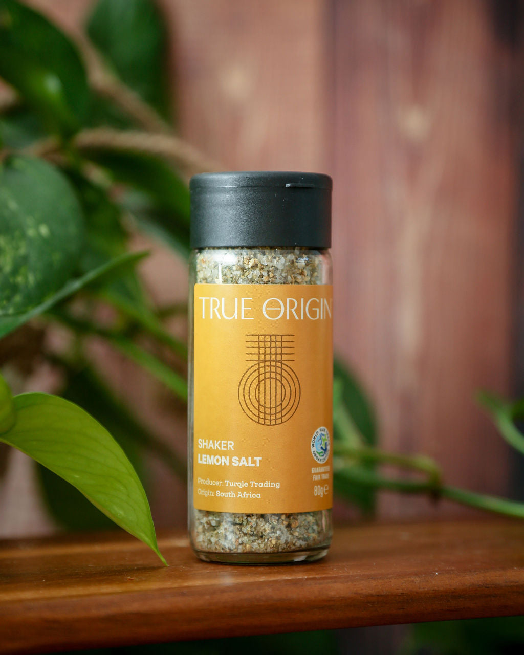 A yellow glass jar of lemon salt with a black lid on a chopping board with some green leaves in the background.